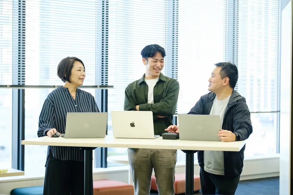 ノートパソコンを置いたテーブルを囲んで立ち話をする社員