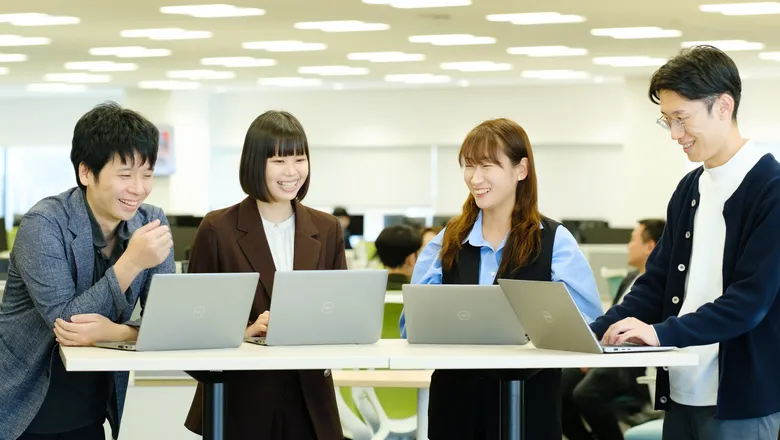 オフィスのテーブルでノートパソコンを開き、笑顔で話す社員4人