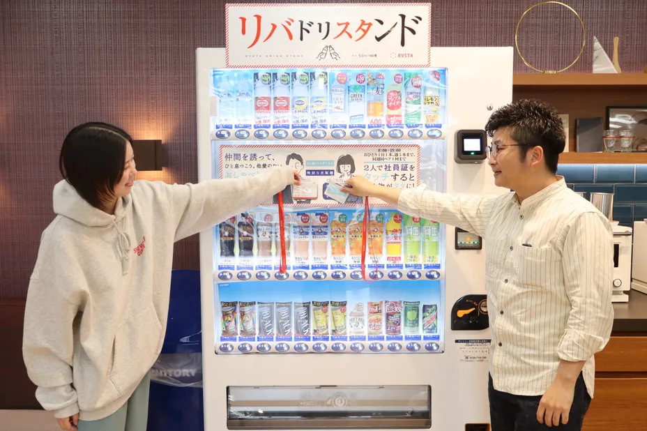 社員証をタッチして使う自動販売機「リバドリ」の前に立つ2人の社員