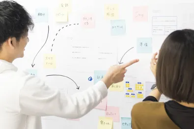 付箋の貼られたホワイトボードを指しながら話し合う社員