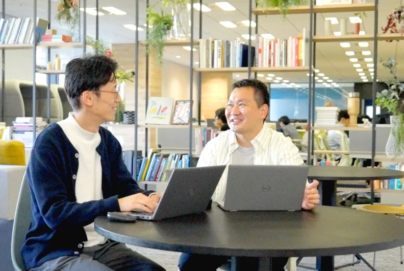 丸テーブルでノートパソコンを開いて話す2人の社員