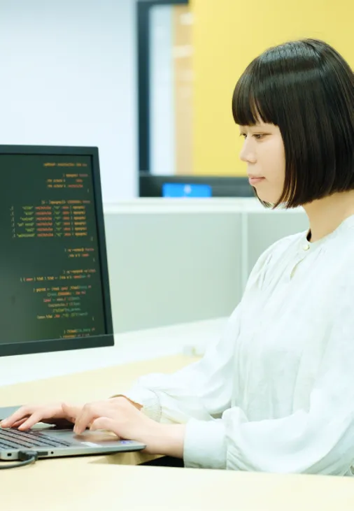 プログラムのコードが映るモニターの横でノートパソコンを操作する社員