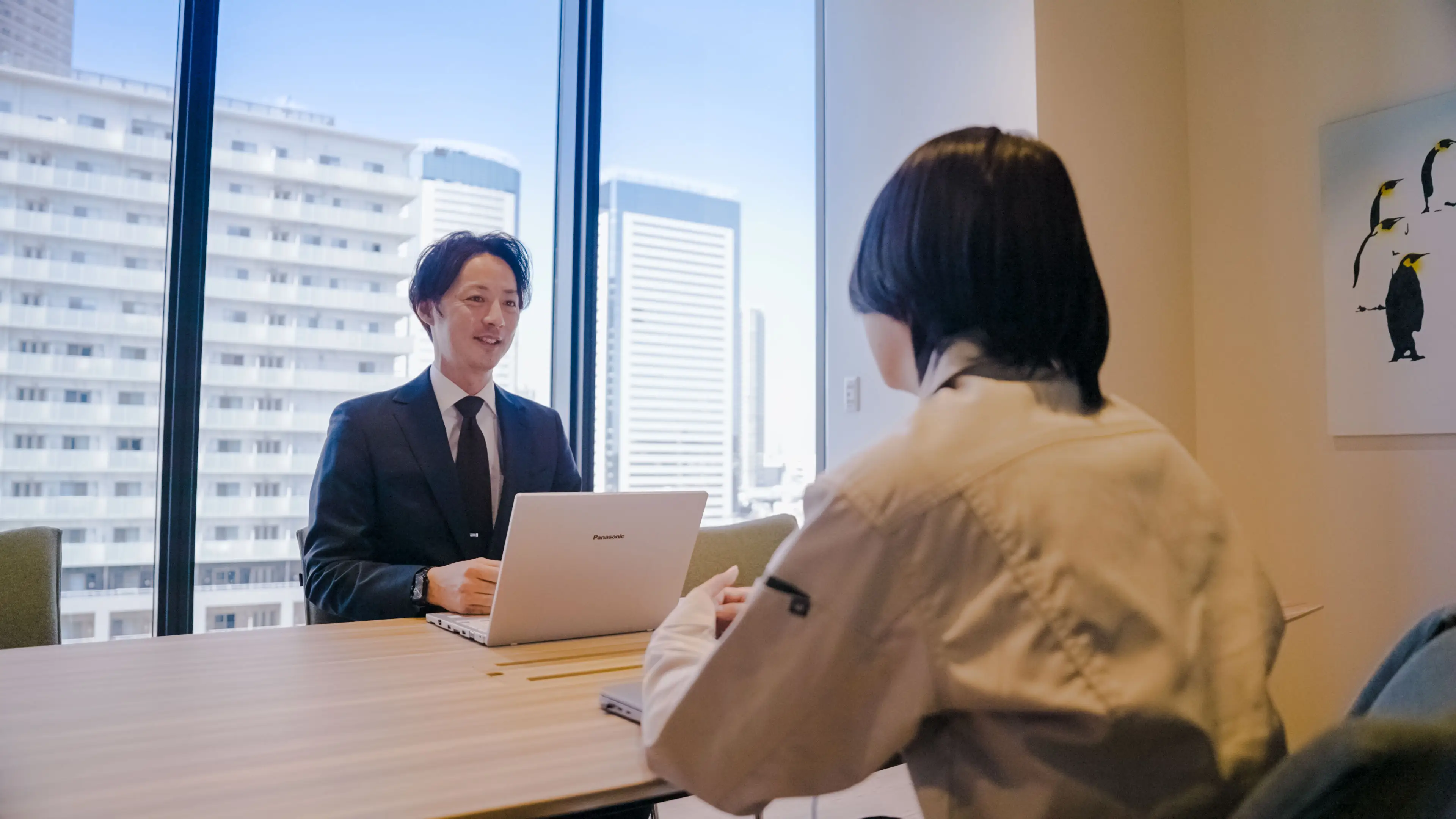 ノートパソコンを前に窓際で打ち合わせをするスーツ姿の社員