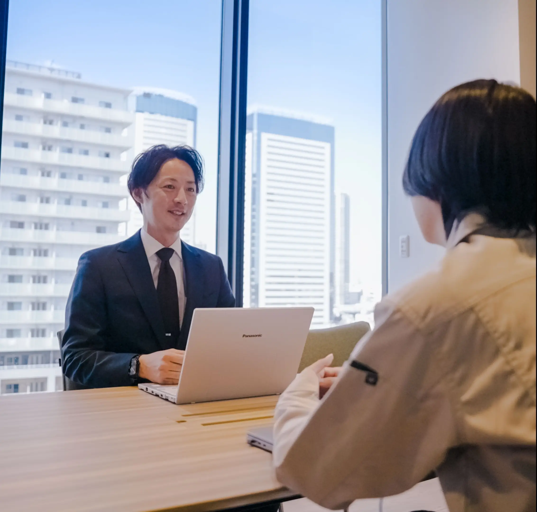 ノートパソコンを前に窓際で打ち合わせをするスーツ姿の社員